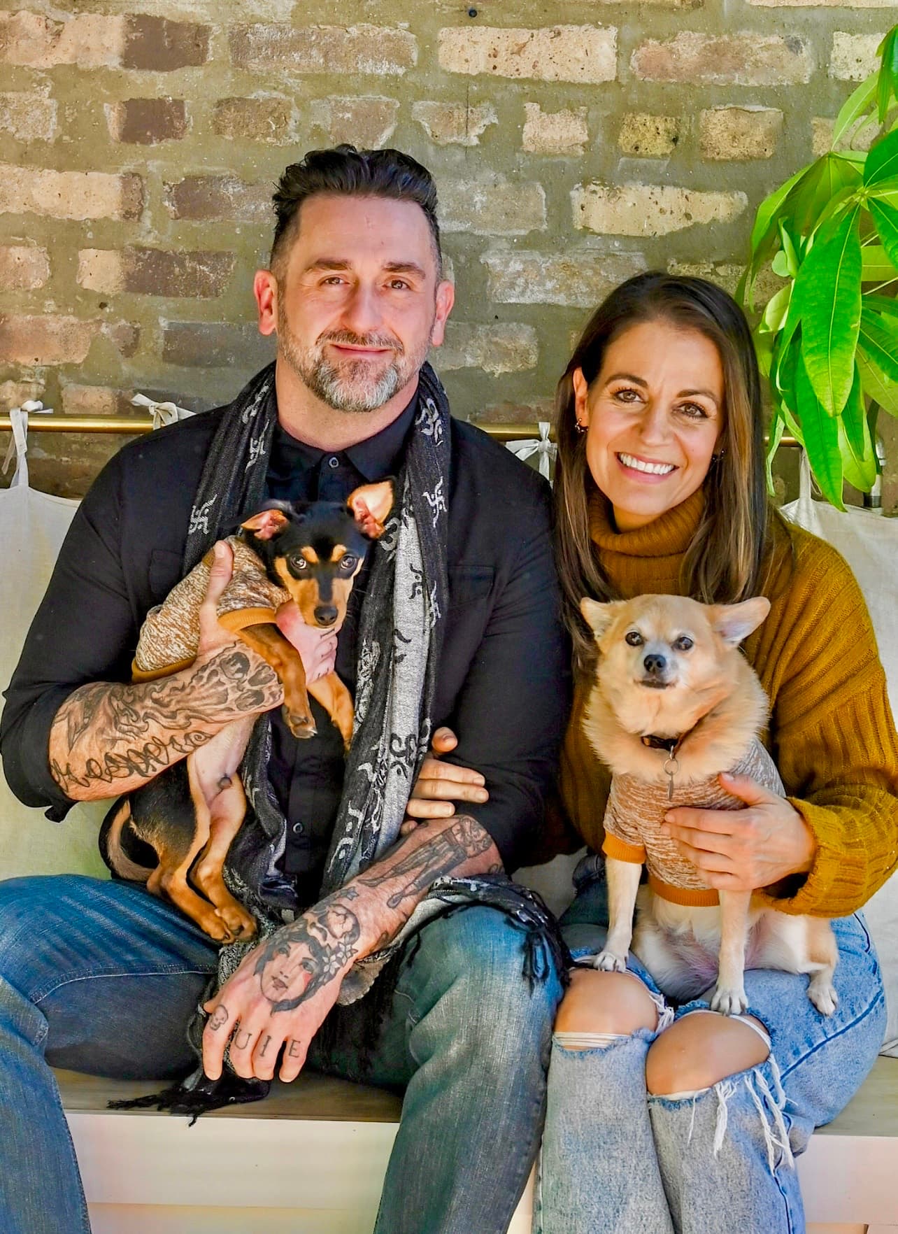 Jenn Dieas and Shane Schwartz seated against weathered brick, each holding one of their dogs